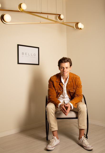 Tiago Curioni sentado sob o pendente linear da Bello, com o quadro da coleção na parede.